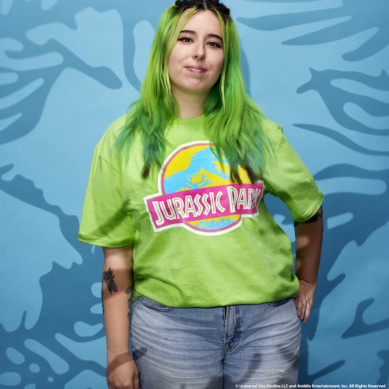 Woman against a blue background wearing a neon t-shirt with the Jurassic Park logo on it